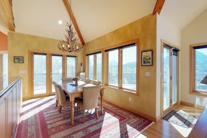 A spacious dining area with a wooden table and chairs, large windows, and a chandelier. - Luxury Home with Upscale Fixtures, WiFi, Private Hot Tub, & Mountain Views (Silverthorne)