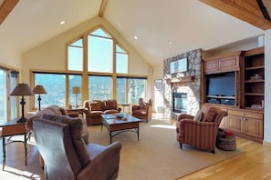 A spacious living room with a high ceiling, large windows, a stone fireplace, and wooden cabinetry. - Luxury Home with Upscale Fixtures, WiFi, Private Hot Tub, & Mountain Views (Silverthorne)