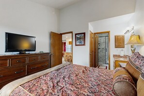 A bedroom with a bed, a television on a dresser, a bathroom with a shower, and a door leading to another room. - Charming Mountain View Condo with WiFi, Fireplace (Silverthorne)