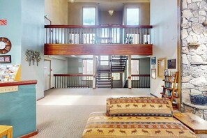A spacious room with a high ceiling, a staircase leading to an upper level, and a bookshelf filled with books. - Charming Mountain View Condo with WiFi, Fireplace (Silverthorne)