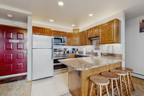 A kitchen with wooden cabinets, a stainless steel refrigerator, a microwave, an oven, and a granite countertop with bar stools. - Charming Mountain View Condo with WiFi, Fireplace (Silverthorne)
