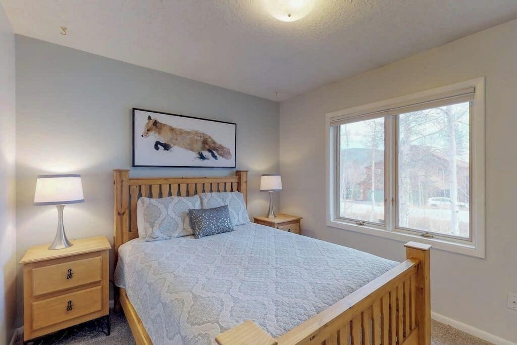 A bedroom with a wooden bed, two bedside tables with lamps, a framed picture on the wall, and a window with a view.