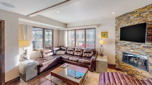 A modern living room with a leather sectional sofa, a coffee table, a TV mounted on a stone wall, and large windows. - 1 Block to Vail Mountain! Village Condo w/ Fireplace, Great Views & Shared Pool (Vail)