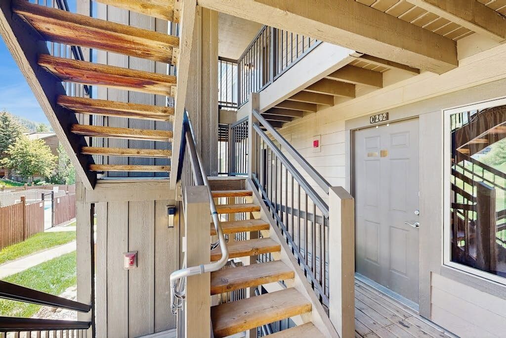 Un escalier en bois à plusieurs niveaux, doté de rampes en métal, une porte portant l’inscription « C202 », et une vue sur un quartier résidentiel.