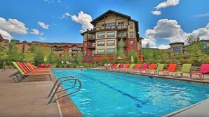 A swimming pool with lounge chairs, a multi-story building with balconies, and a clear blue sky. - Family-friendly condo w/mountain & valley views, WiFi, shared pool, hot tub (Granby)