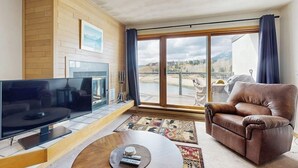 Una sala de estar con televisión de pantalla plana, sofá de cuero y un ventanal con vista a un cuerpo de agua y montañas.