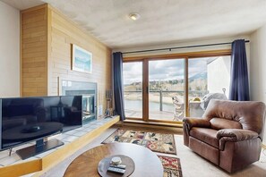 A living room with a flat-screen TV, leather sofa, and a large window with a view of a body of water and mountains. - Lakefront retreat with mountain views, covered parking & updated interiors (Dillon)