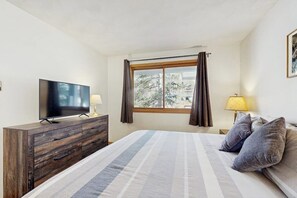 A bedroom with a bed, a TV on a wooden dresser, a window with curtains, and a lamp on a bedside table. - Lakefront retreat with mountain views, covered parking & updated interiors (Dillon)