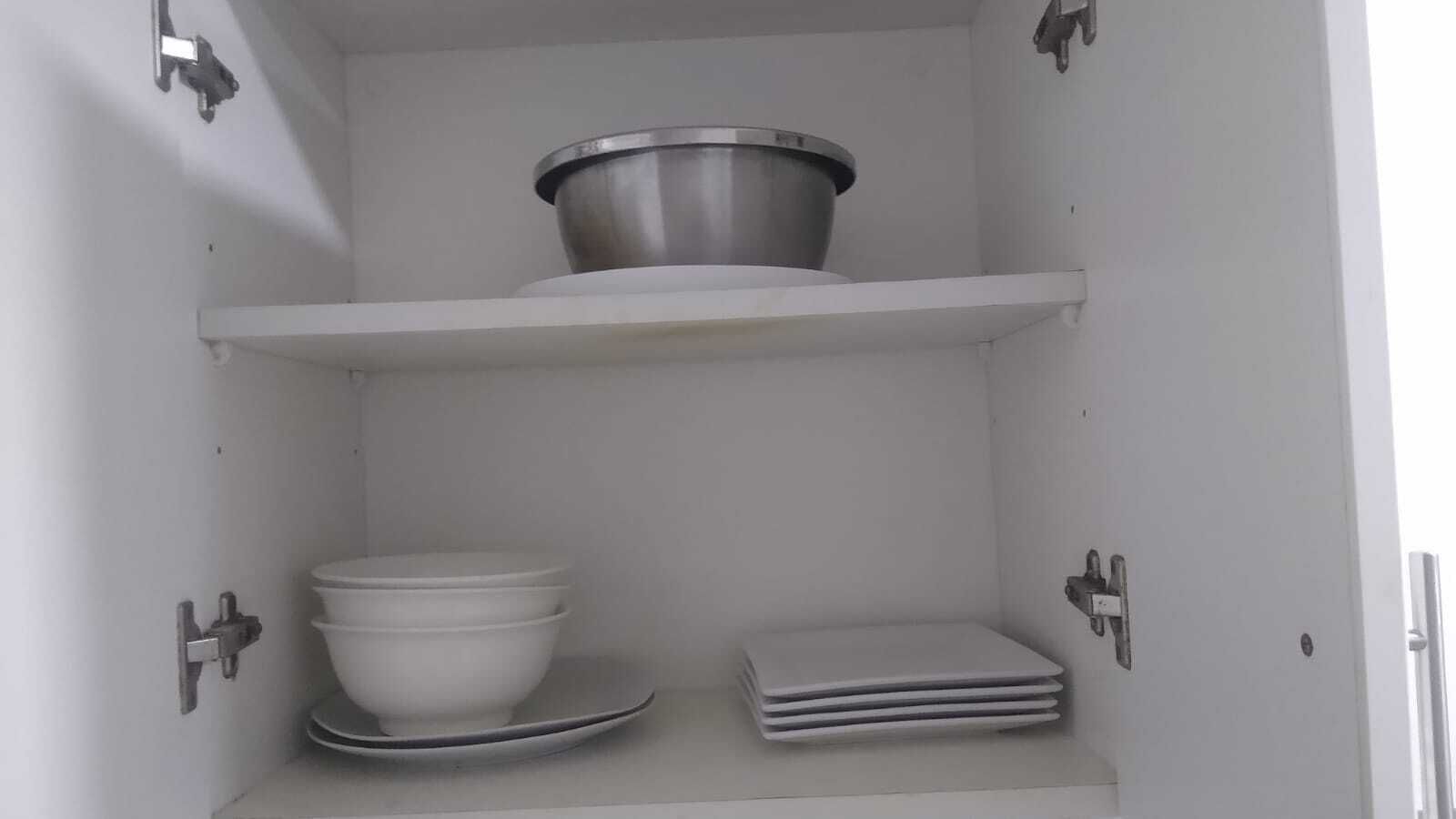 A kitchen cabinet with a stainless steel pot on the top shelf, a white bowl on the middle shelf, and stacked white plates on the bottom shelf.