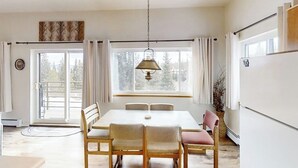 Un coin repas comprenant une table et des chaises, un luminaire suspendu, une fenêtre avec des rideaux et une vue sur les arbres à l’extérieur.