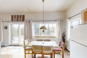 A dining area with a table and chairs, a hanging light fixture, a window with curtains, and a view of trees outside. - Mountaintop condo with private balcony (Silverthorne)