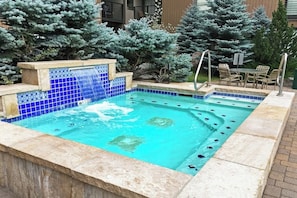 Ein rechteckiger Swimmingpool mit blauem Mosaikrand, einer kleinen Wasserfallanlage und einem Sitzbereich mit Stühlen und einem Tisch.