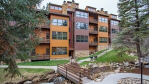 A multi-story building with balconies, a stone facade, and a landscaped area with a stream and wooden bridge. - Kutuk 108 Ski, stay, & play - Steamboat Springs getaway only steps away from the mountain (Steamboat Springs)
