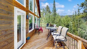 Una terraza de madera con una mesa y sillas, un ventanal de vidrio y vistas a árboles y colinas.