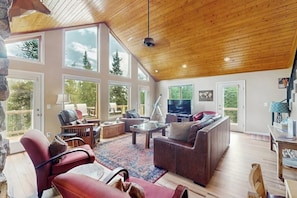 A spacious living room with a large TV, comfortable seating, and large windows offering a view of the outdoors. - Secluded Mountain Cabin w/ Hot Tub, Fireplace & Stargazing Deck near Breck (Alma)