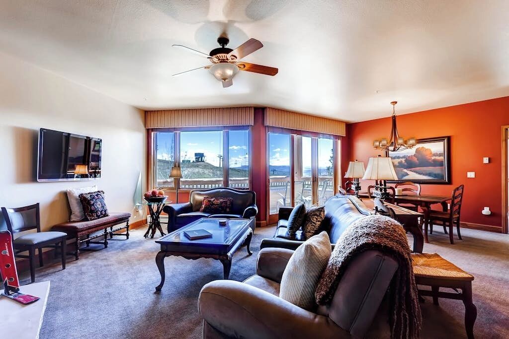 A living room with a large window, a ceiling fan, a flat-screen TV, and a dining area with a table and chairs.