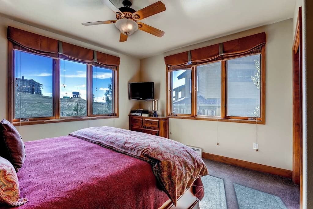 Ein Schlafzimmer mit einem Bett, Deckenventilator, Fernseher und großen Fenstern mit Vorhängen.