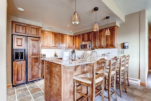 A kitchen with wooden cabinets, a central island with bar stools, a microwave, and a refrigerator. - Luxury Ski-In/Out Condo w/Free WiFi, Fireplace, W/D, Shared Pool, Hot Tub, Grill (Steamboat Springs)