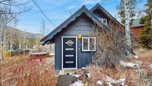 Une petite maison en bois, de couleur sombre, avec un toit à pignon, une porte d’entrée ornée d’une guirlande et un petit porche.