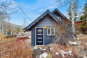 A small, dark-colored wooden house with a gabled roof, a front door with a wreath, and a small porch area.