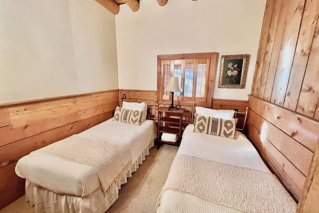A room with two single beds, wooden walls, a window with blinds, a small table, and a picture on the wall.