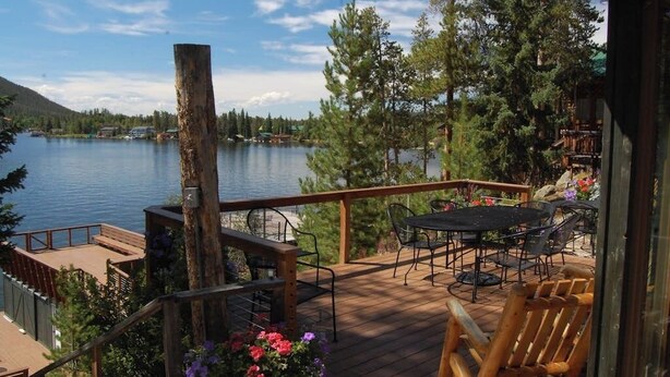 A wooden deck with outdoor furniture, overlooking a lake and surrounded by trees. - Original charmer with expansive waterfront deck, dock, fireplace - walkable area (Grand Lake)
