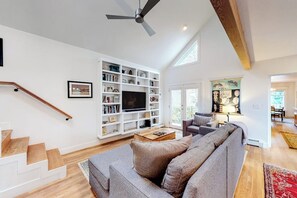 Ein Wohnzimmer mit grauem Sofa, einem Fernseher auf einem weißen Regal und einer Treppe mit Holzdielen.