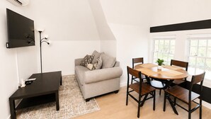 A modern living room with a sofa, a dining table with chairs, a TV mounted on the wall, and a large window.