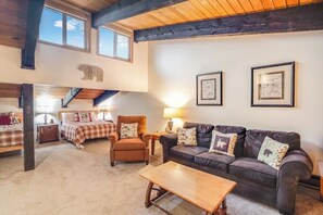Ein gemütliches Wohnzimmer mit einer Couch, einem Sessel und einem Holztisch. An den Wänden befinden sich gerahmte Bilder und eine Bär-Dekoration über der Couch.