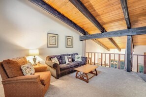 Ein gemütliches Wohnzimmer mit einer Couch, einem Sessel und einem Couchtisch, Holzbalken an der Decke und einem Balkon mit Geländer.