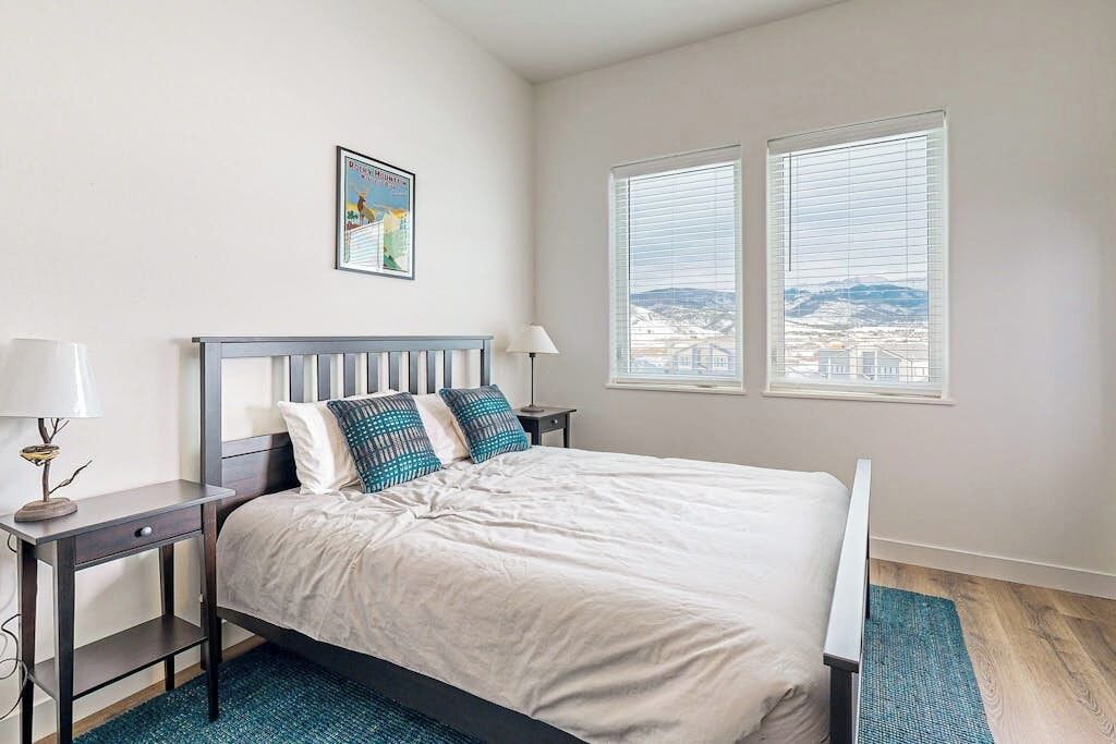 A bedroom with a bed, two bedside tables, a lamp, a framed picture, and a window with a view.