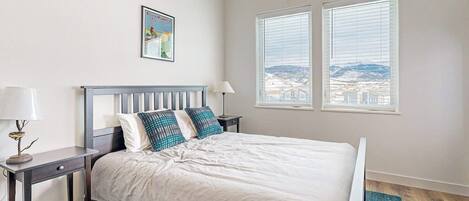 A bedroom with a bed, two bedside tables, a lamp, a framed picture, and a window with a view.