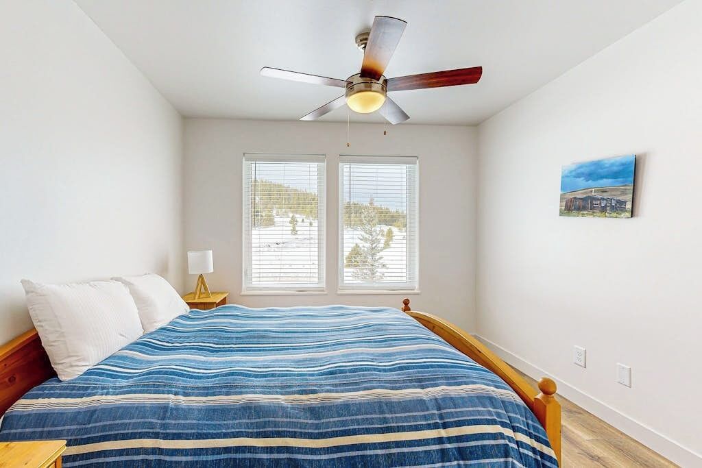 A bedroom with a bed, a ceiling fan, two windows, a nightstand, and a wall-mounted lamp.