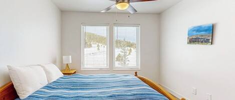A bedroom with a bed, a ceiling fan, two windows, a nightstand, and a wall-mounted lamp.