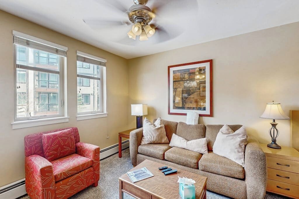 A living room with a red patterned armchair, a beige sofa, a ceiling fan, and a framed artwork on the wall.