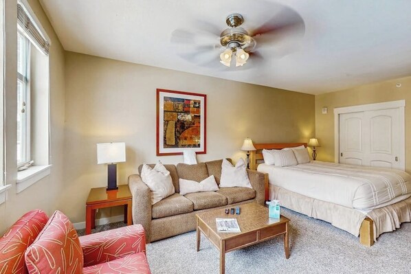 Ein Hotelzimmer mit einem Bett, einem Sofa, einem Couchtisch, einem Deckenventilator, einem gerahmten Bild an der Wand und einem Fenster.