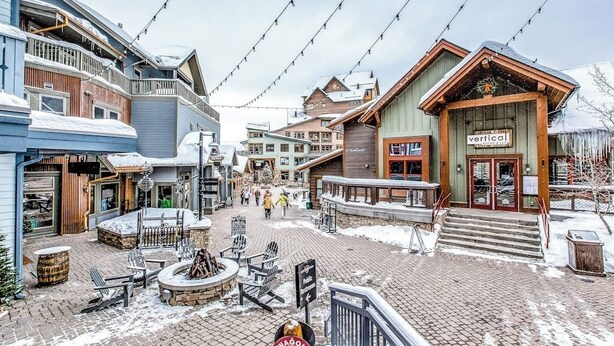 A snow-covered town square with buildings, outdoor seating, and a sign for 'vertical.'