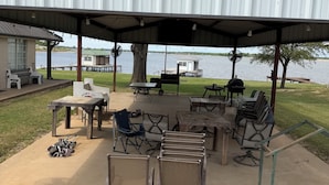 Un área de estar al aire libre con techo, mesas y sillas, una parrilla y vistas a un lago.
