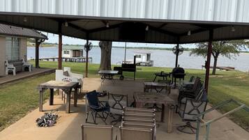 A covered outdoor seating area with tables and chairs, a grill, and a view of a lake.