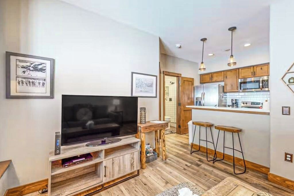 A modern living room with a flat-screen TV, wooden flooring, a kitchen with bar stools, and pendant lighting.