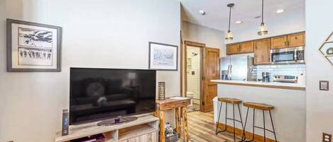 A modern living room with a flat-screen TV, wooden flooring, a kitchen with bar stools, and pendant lighting.