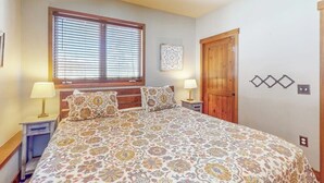 Une chambre à coucher avec un lit, deux tables de chevet avec des lampes, une fenêtre avec des stores, une porte en bois et un ventilateur de plafond.