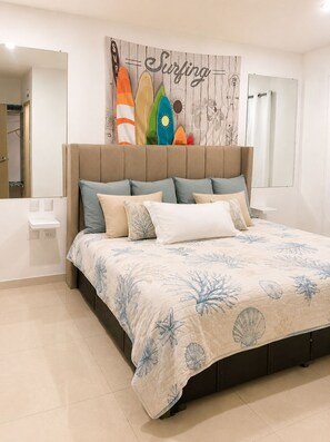 A bedroom with a bed, a nightstand, a mirror, and a wall with a surf-themed artwork. - All-Inclusive Mazatlán Condo for Retirees • Pool • Cleaning • Near Malecón (Mazatlán)