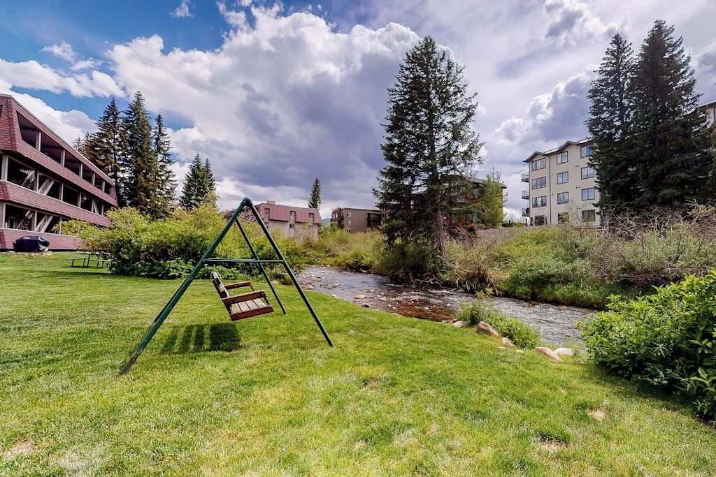A grassy area with a swing set, a stream, and buildings in the background.