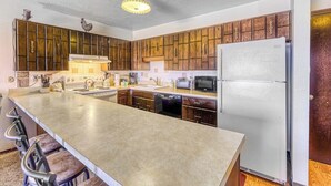 A kitchen with wooden cabinets, a stainless steel refrigerator, a microwave, and a bar stool area. - Mountain view condo w/ a shared pool, hot tub, Ping-Pong, & pool table (Dillon)
