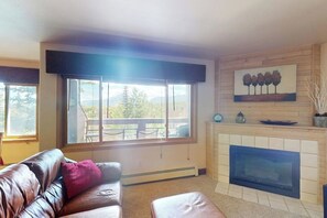 A living room with a leather sofa, a fireplace, a large window with a view, and a painting on the wall. - Wildernest Condo Close to Top Ski Resorts  + Free WiFi (Silverthorne)