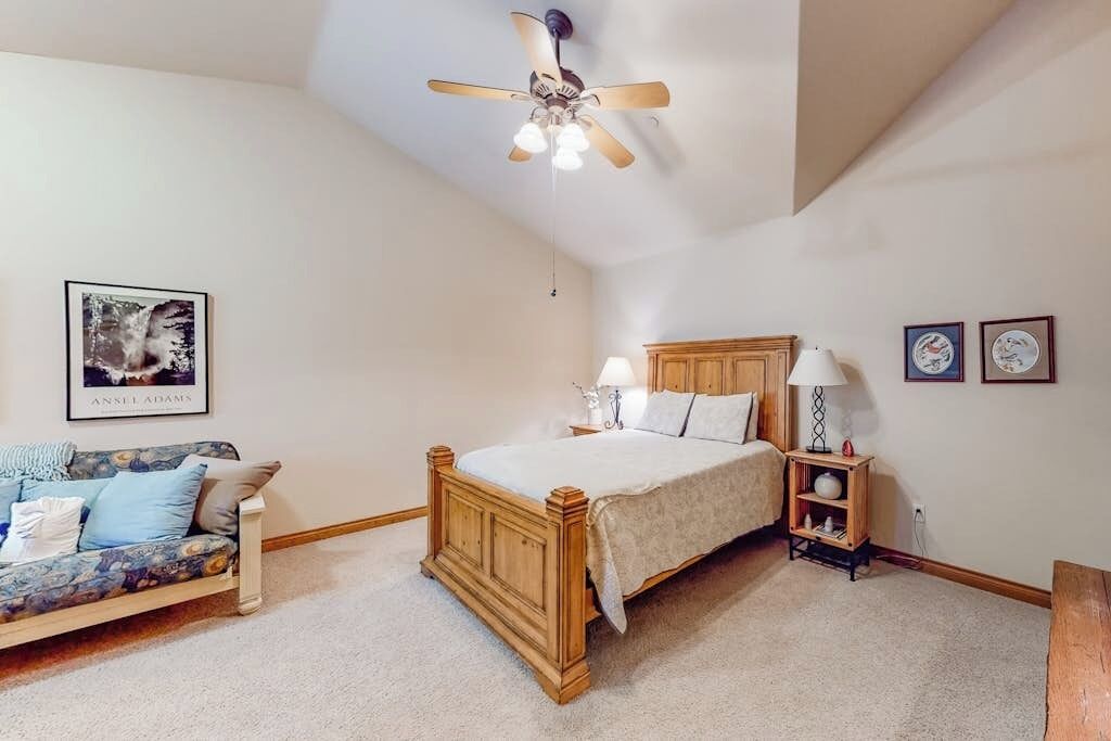A bedroom with a wooden bed, a ceiling fan, a nightstand, a lamp, a sofa, and a framed picture on the wall.