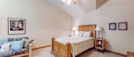 A bedroom with a wooden bed, a ceiling fan, a nightstand, a lamp, a sofa, and a framed picture on the wall.