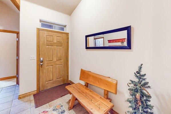 A hallway with a wooden door, a bench, a small Christmas tree, and a framed picture on the wall. - Blue River condo with water-view balcony, walking distance to the outlets (Silverthorne)