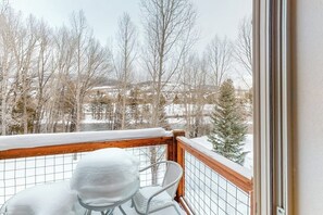 Ein Balkon mit einem Metallstuhl und einem Tisch, der mit Schnee bedeckt ist, mit Blick auf eine verschneite Landschaft mit Bäumen und Gebäuden.